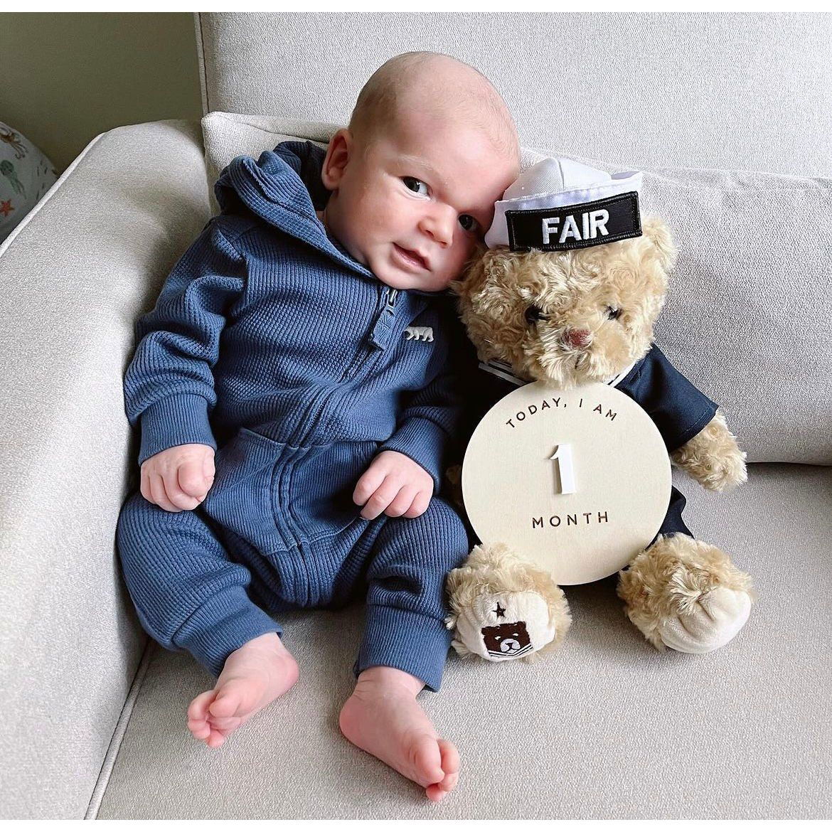 Baby in a blue outfit sitting next to a teddy bear with a 'Today I am 1 month' sign, wearing a 'FAIR' headband.
