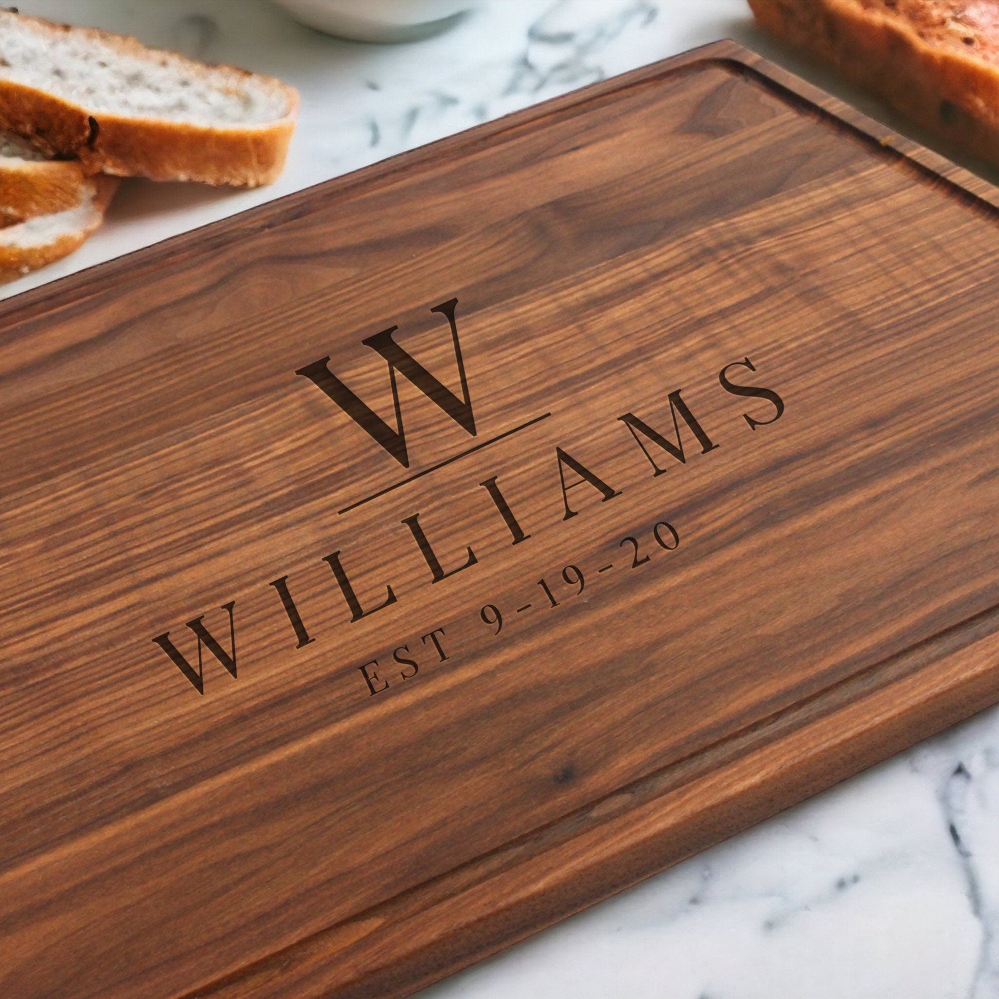 Wooden cutting board with engraved 'WILLIAMS' and 'EST 9-19-20' on a marble surface with bread slices.